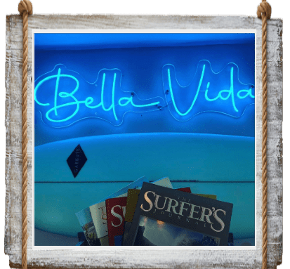 Neon sign reading 'Bella Vida' above vintage surf magazines.